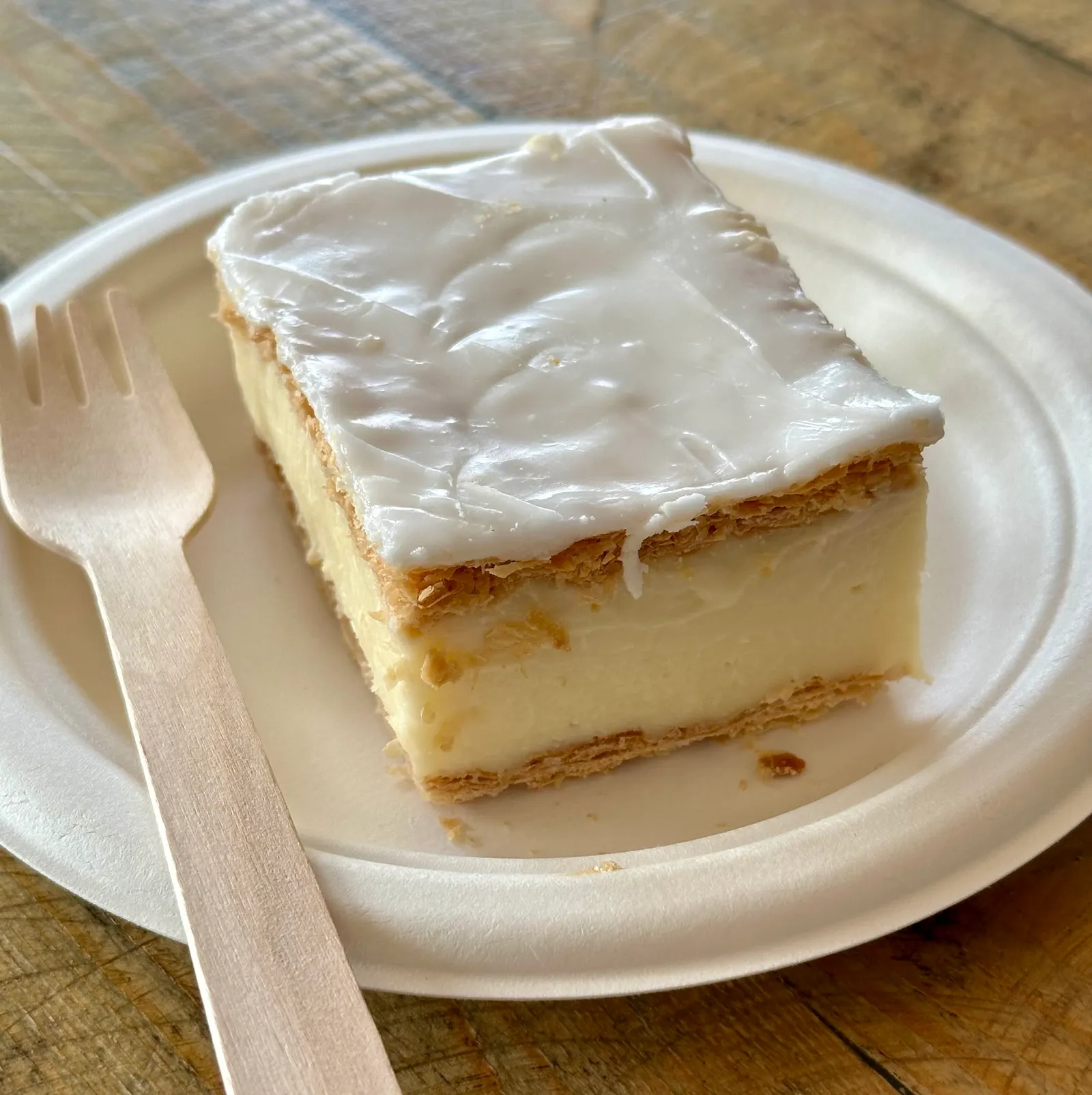 Vanilla slice with white icing and custard on a paper plate with a wooden fork