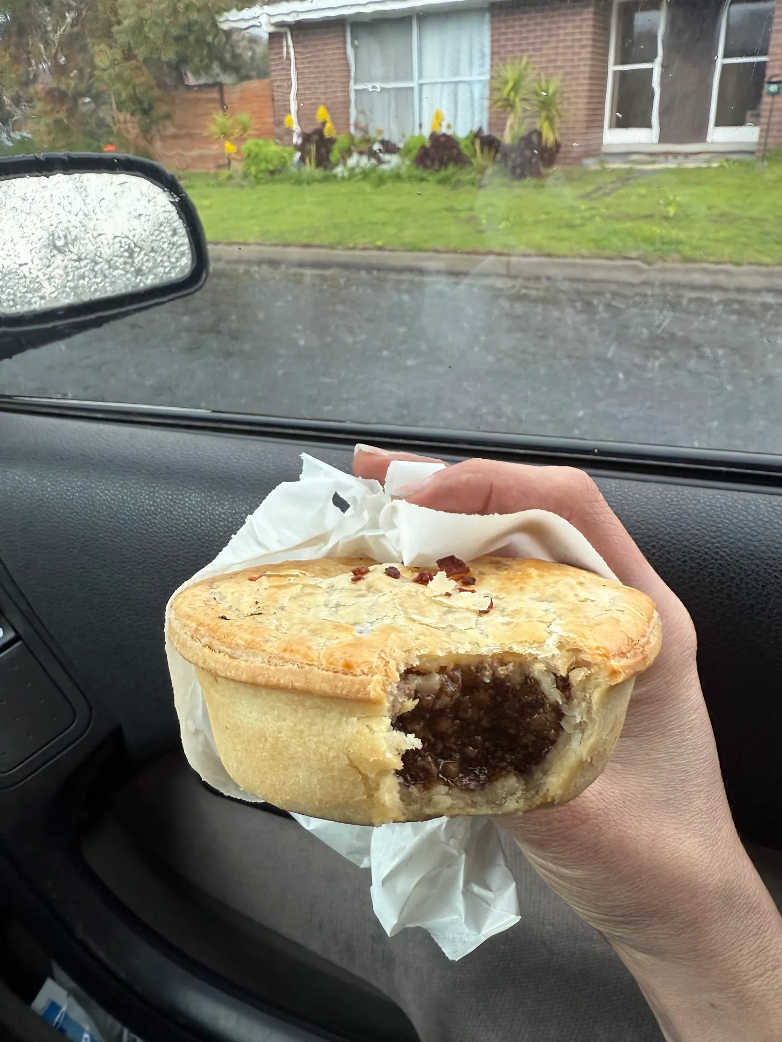 Hand holding a bitten golden meat pie in paper inside a car on a rainy day
